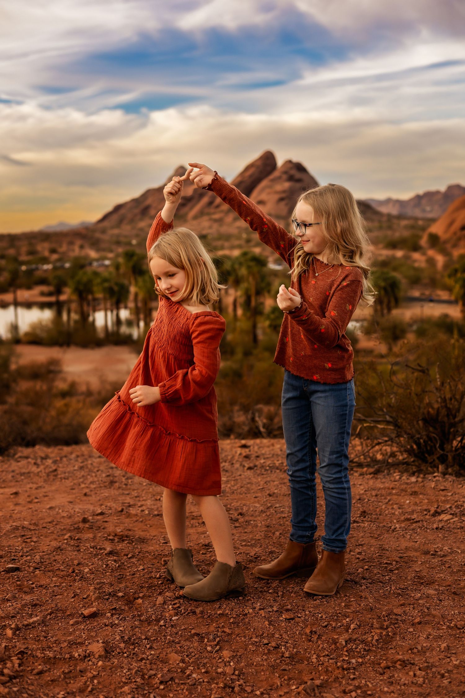 Young siblings in red and denim outfits explore desert terrain during golden hour with mountains and palm trees in background.