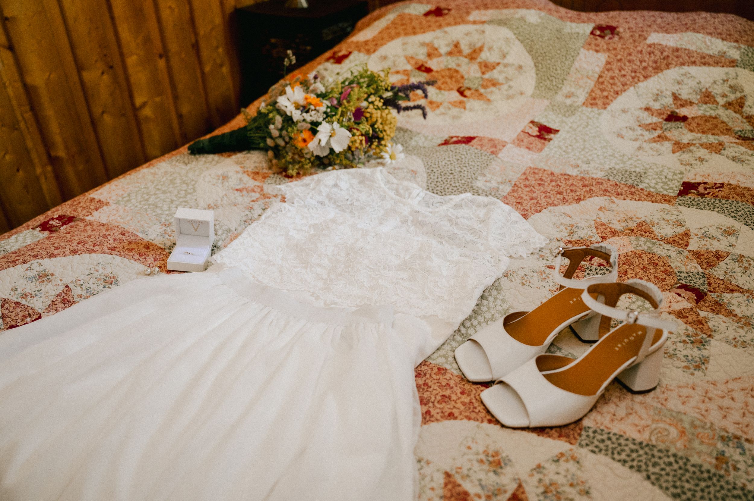 A white wedding dress and brown sandals laid out on a floral patterned bedspread with a bouquet of flowers.
