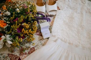 White wedding dress, bridal shoes, and colorful wildflower bouquet arranged on vintage floral pattern textile.