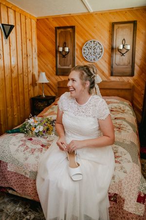 A white lace wedding dress sits against wood paneled walls decorated with vintage decor and a floral patterned quilt.