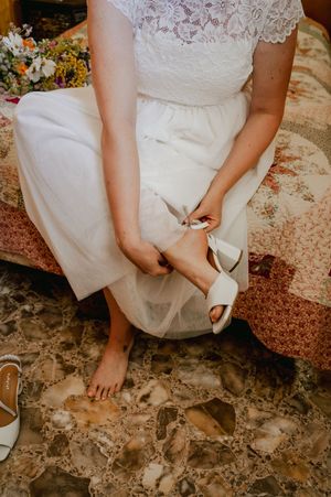 A close-up of a bride in a white lace dress adjusting her white sandal while sitting on a textured stone surface.