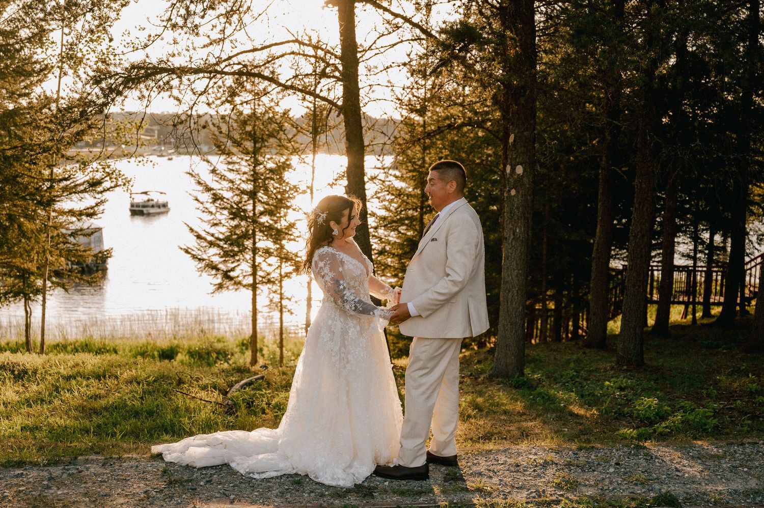 Two people in wedding attire walk hand in hand across a sunlit lawn with evergreen trees in the background.