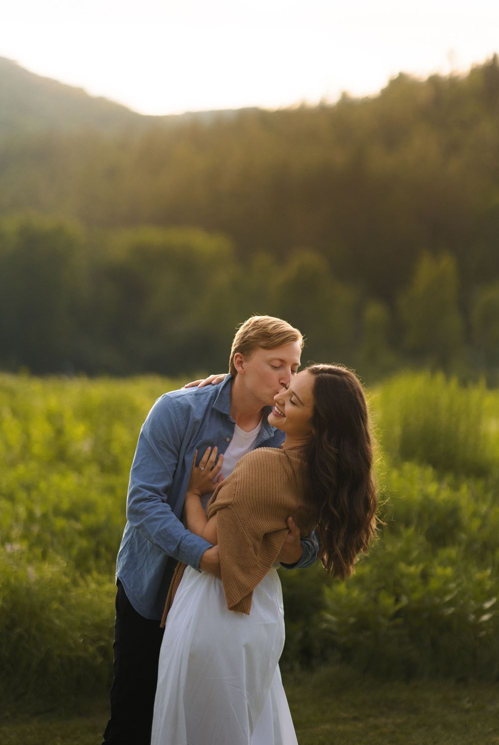 A romantic couple shares an intimate embrace in a sunlit meadow at golden hour with mountains in the background.