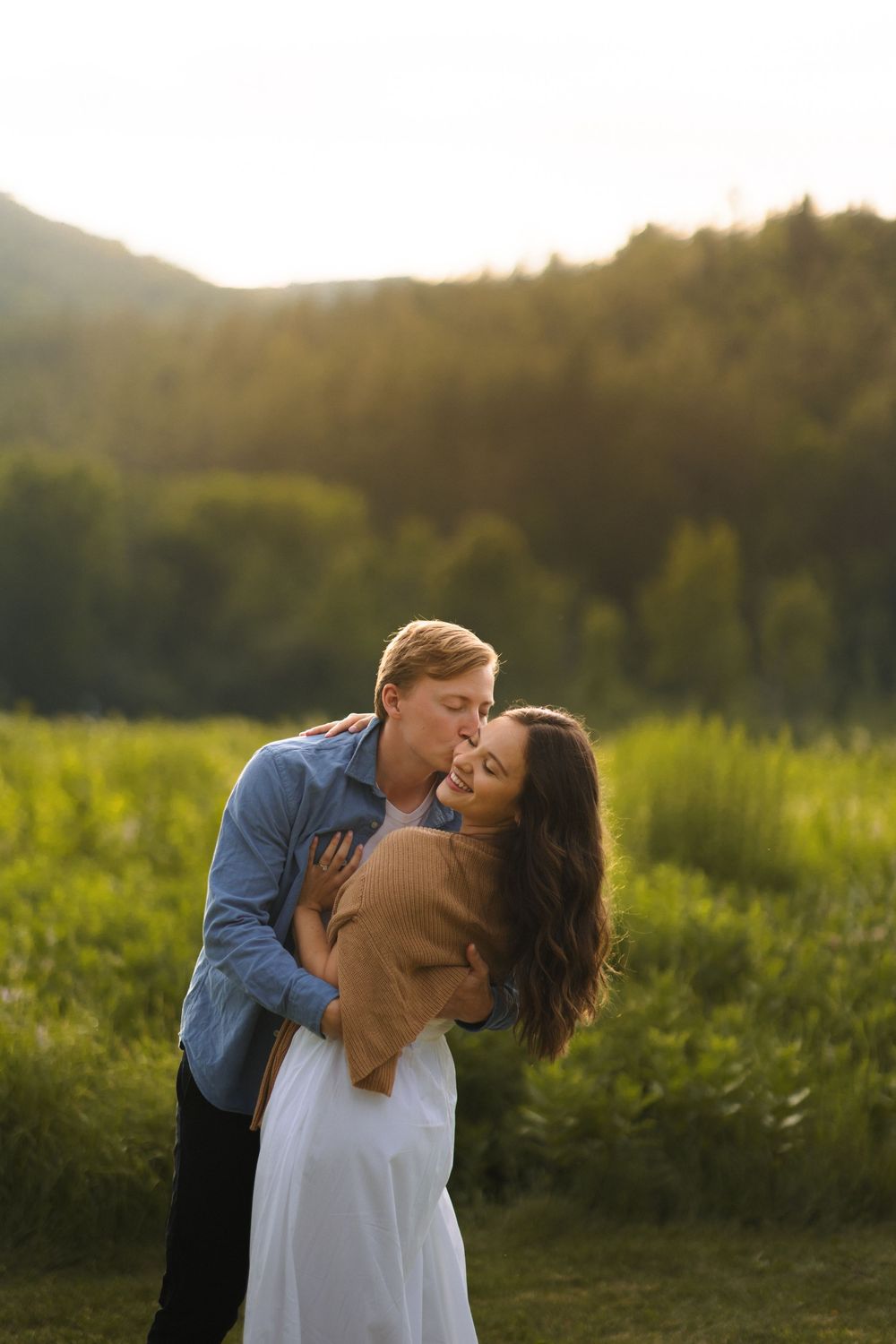 A romantic couple embraces in a sunny meadow with mountain backdrop during golden hour.