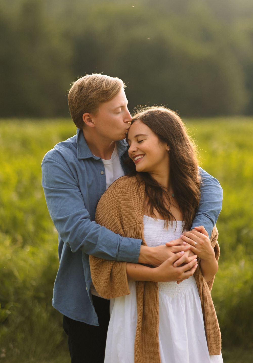 A romantic couple embraces in a sunlit meadow, with her wearing a white dress and tan cardigan.