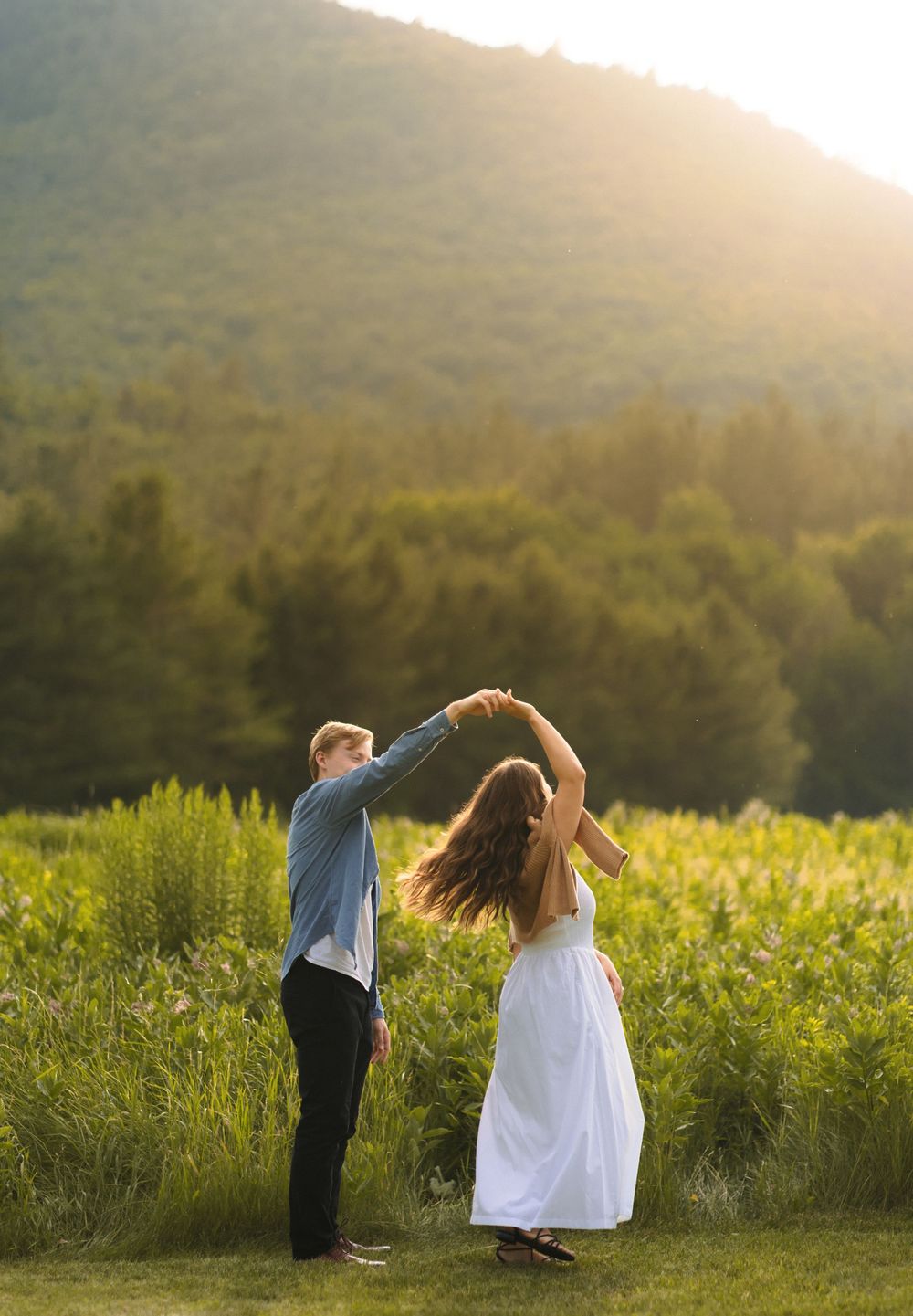 A couple dances together in a sunlit meadow with mountains and forest in the background during golden hour.