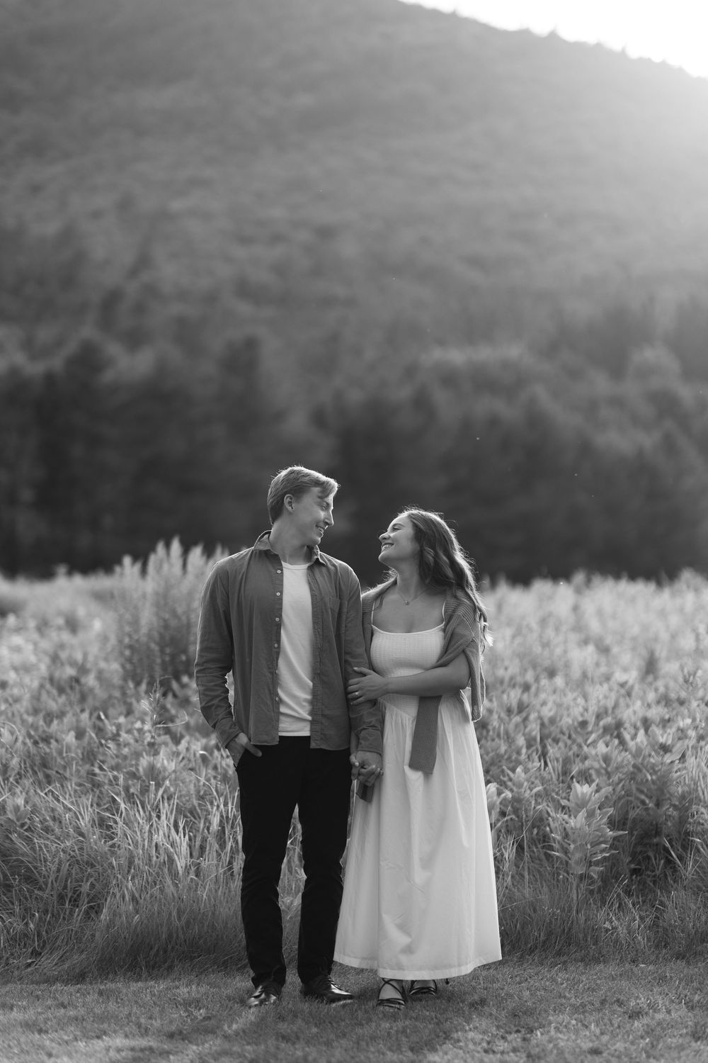 A couple walks together through a misty meadow of tall grasses at sunset against mountain backdrops.