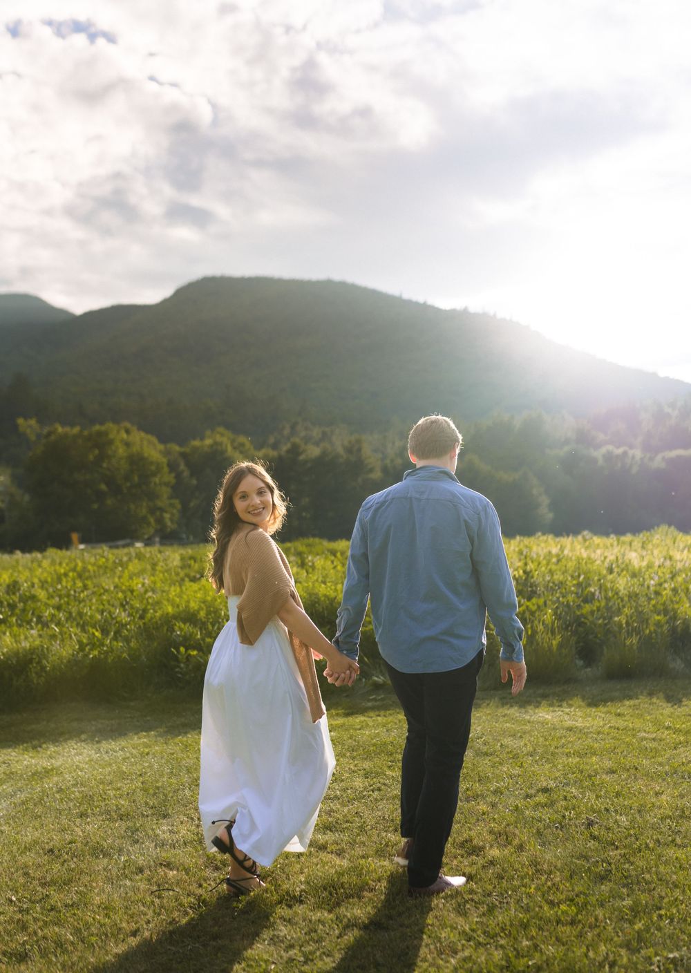 A couple walks hand in hand through a mountain meadow at sunset.