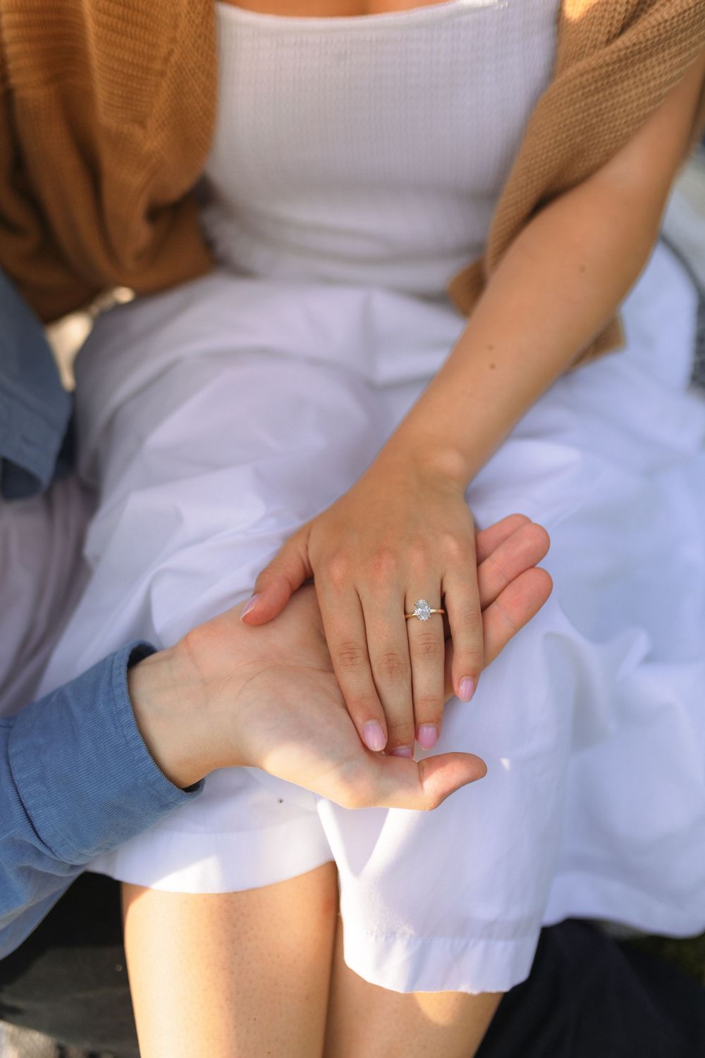 Two hands clasped together showing an engagement ring against a white dress and brown cardigan.