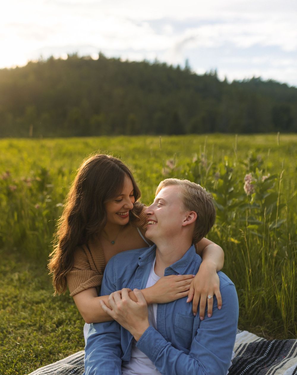 A couple laughs together while cuddling on a blanket in a sunny mountain meadow.