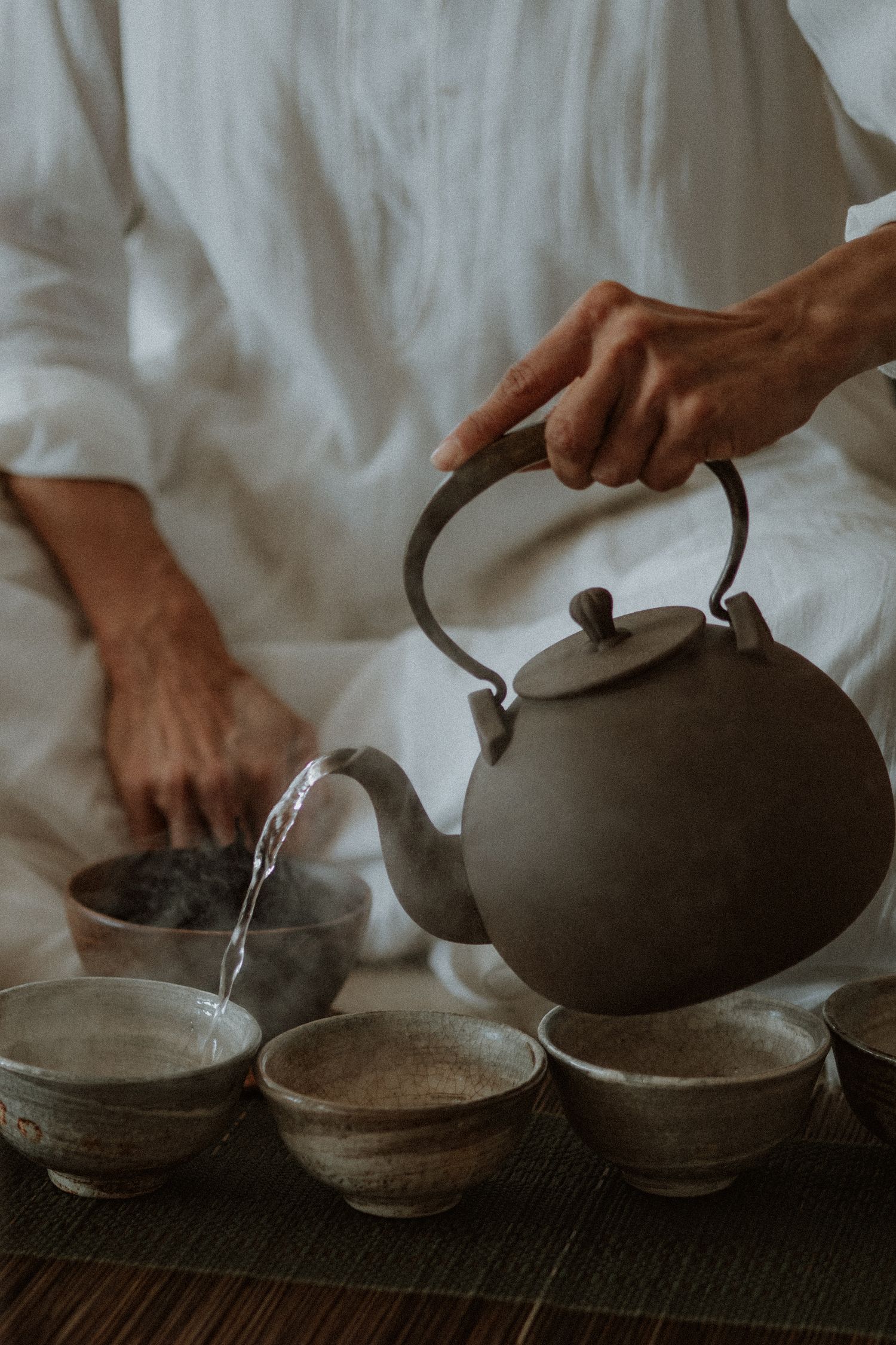 A person pours hot water from a rustic clay teapot into traditional ceramic tea bowls on a bamboo mat.