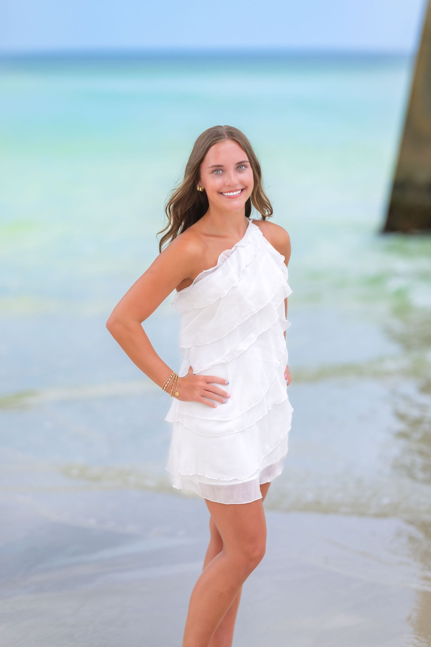 Figure in a flowing white one-shoulder dress poses gracefully by the turquoise ocean on a sunny beach.