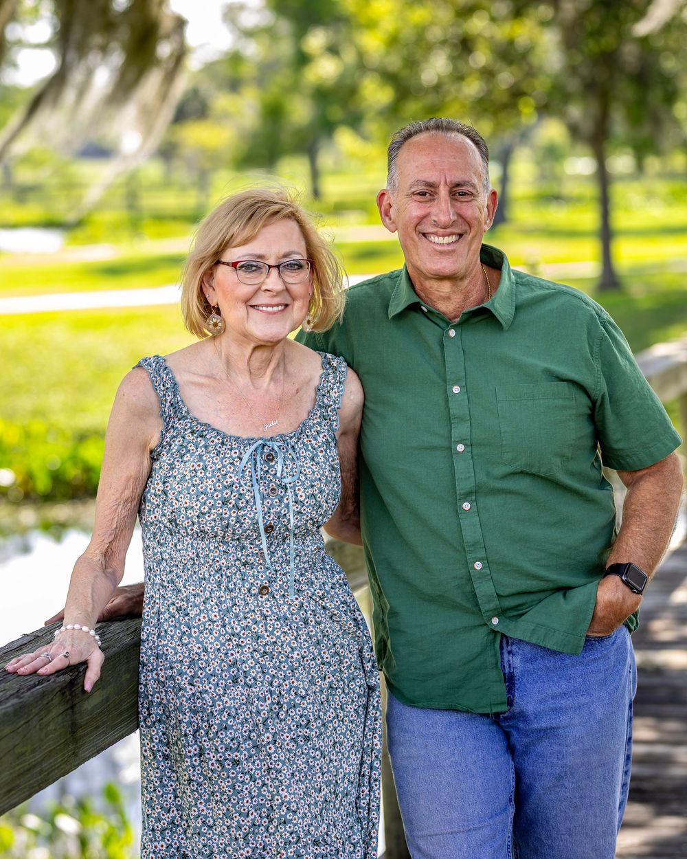 A couple stands outdoors in a sunny park against lush greenery, wearing casual summer attire.