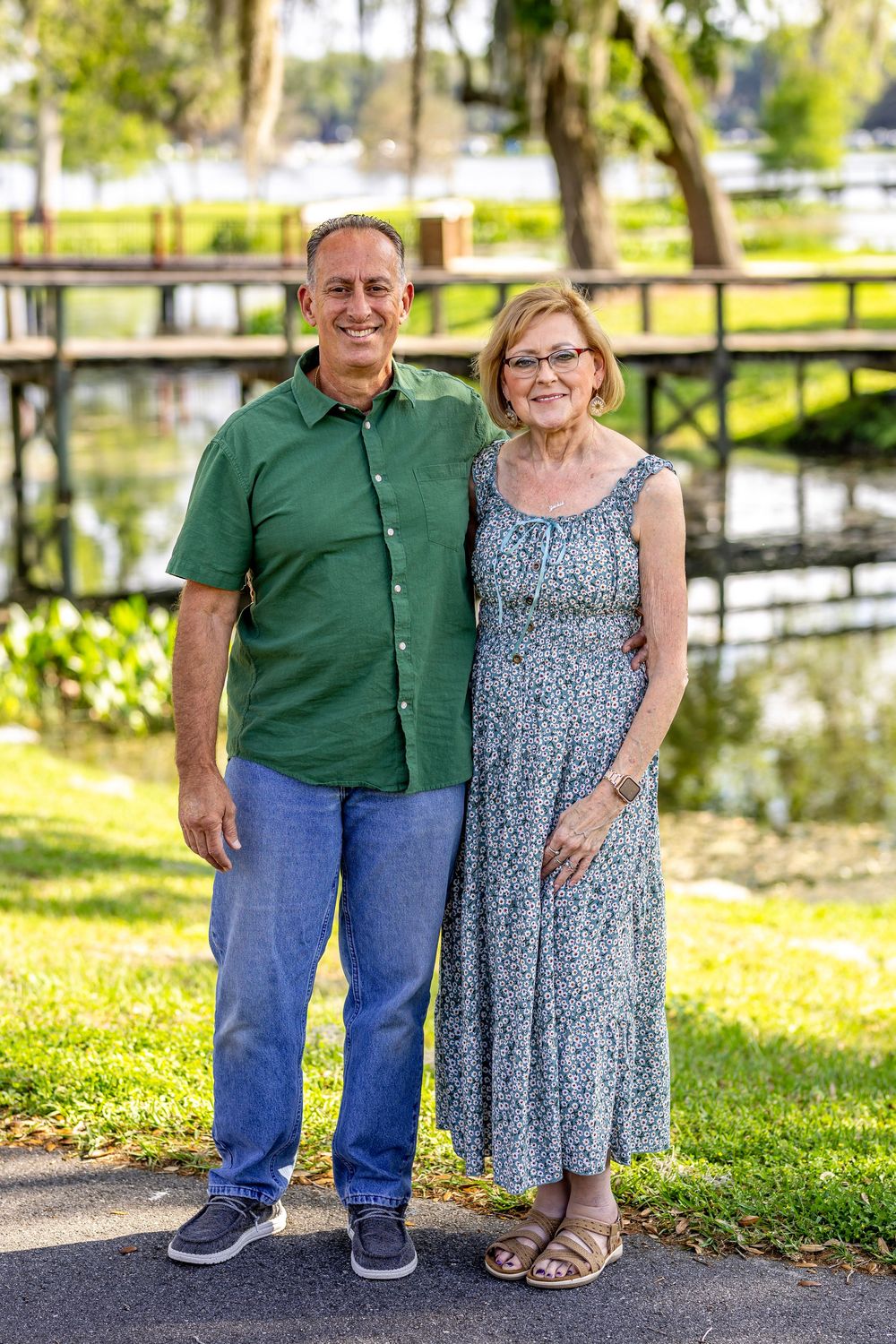 Couple poses together on walkway with lush greenery and waterfront in background.