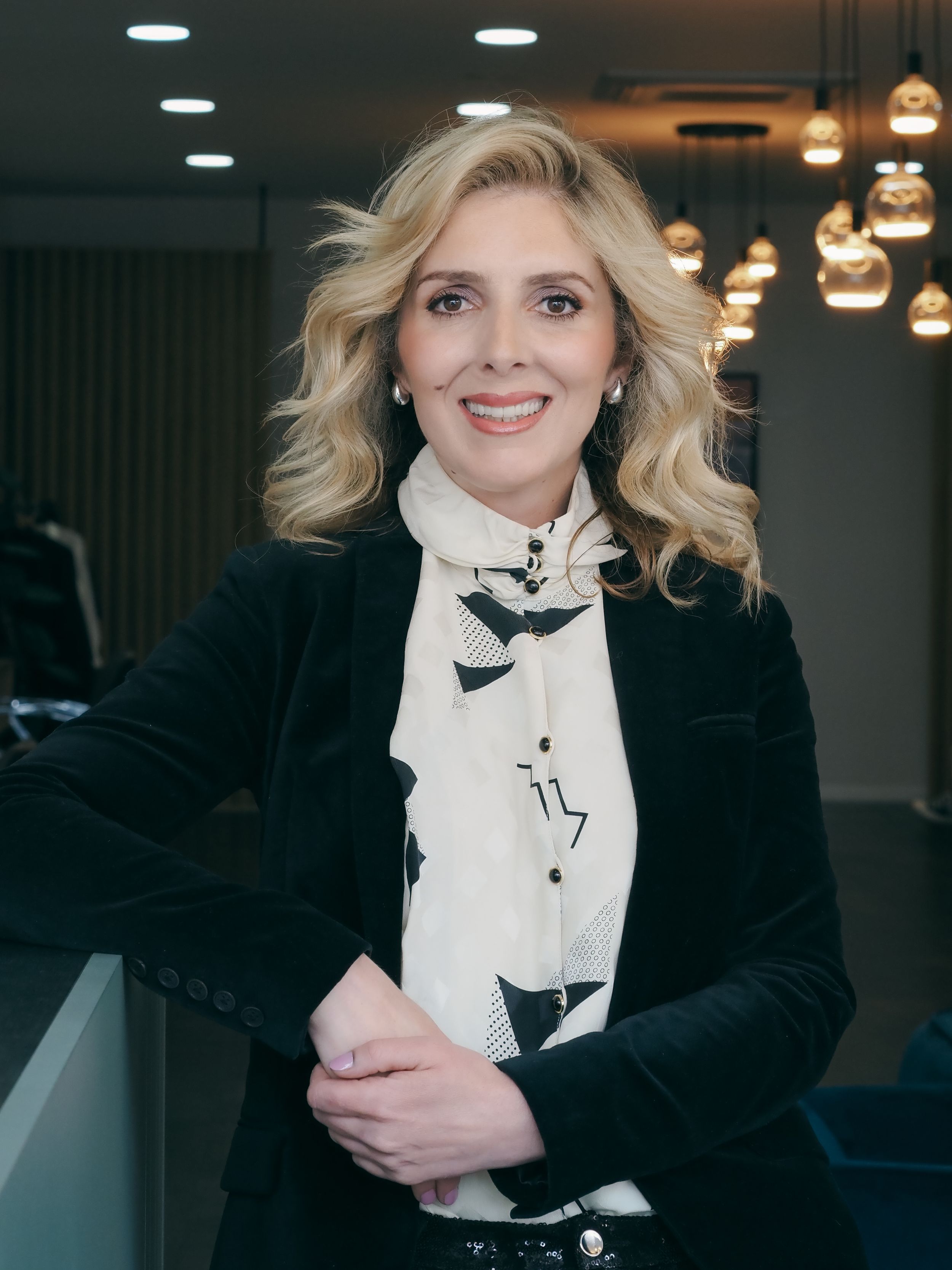 Professional portrait of someone in a black blazer and patterned white blouse against an office interior with pendant lights.