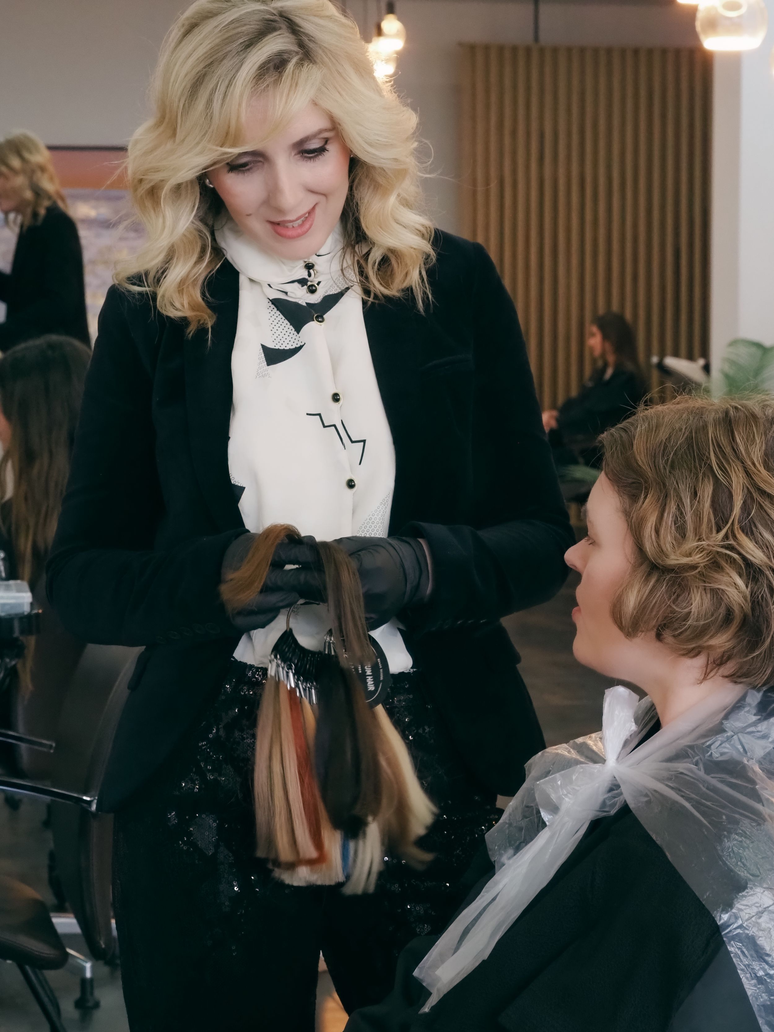 A hairstylist wearing black consults with a client in a salon chair while holding hair tools.