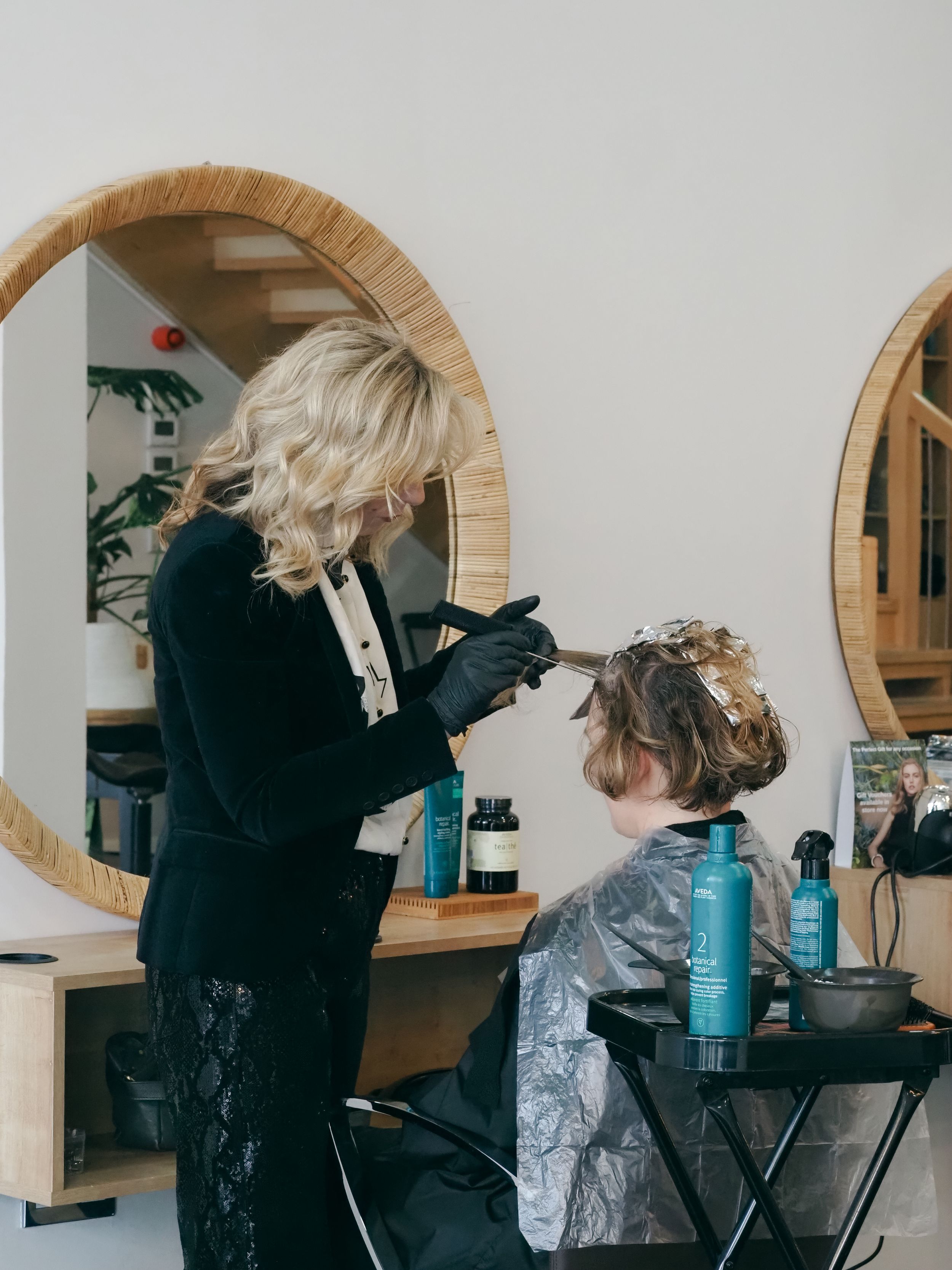 Hairstylist working on a client's hair in front of circular bamboo-framed mirror in modern salon.