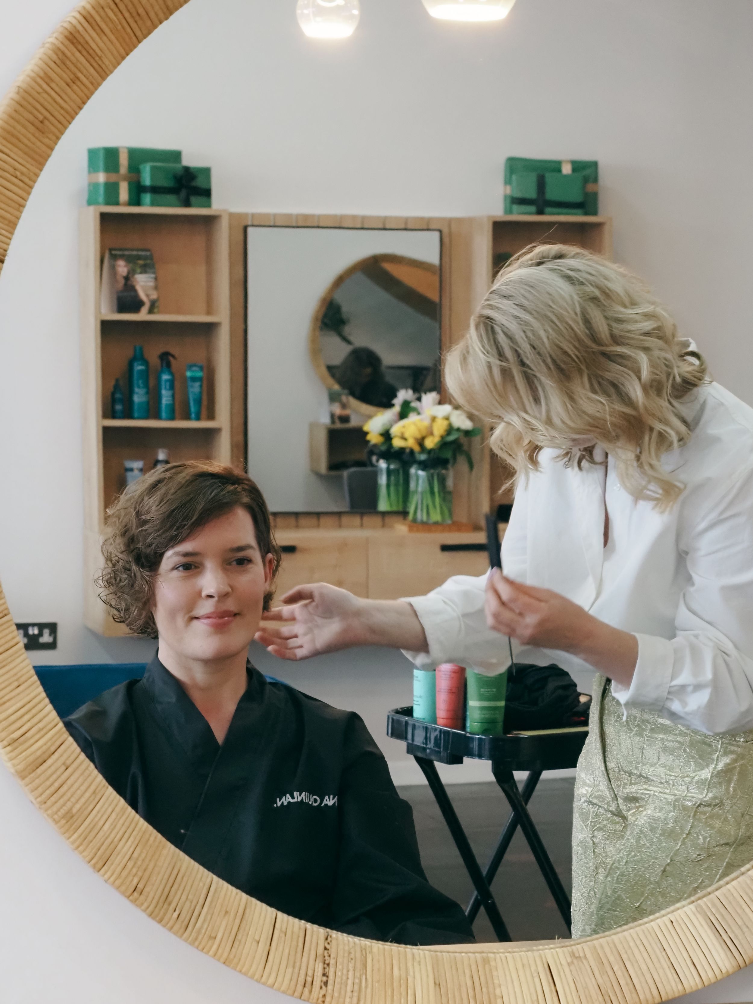 View through round mirror showing salon professional styling hair with products on shelves.