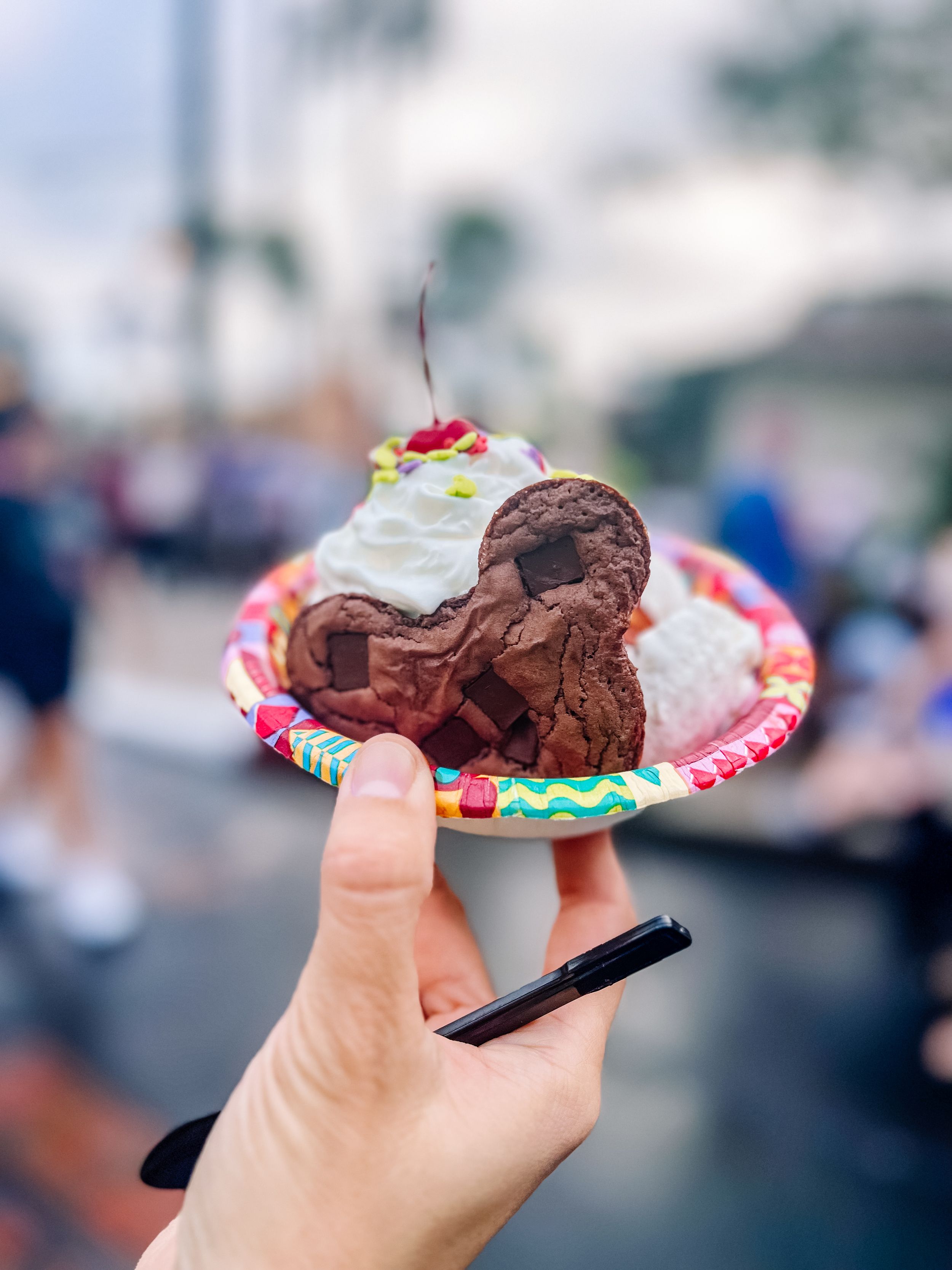 Mickey-shaped chocolate ice cream sundae with whipped cream and cherry on a decorative paper plate.