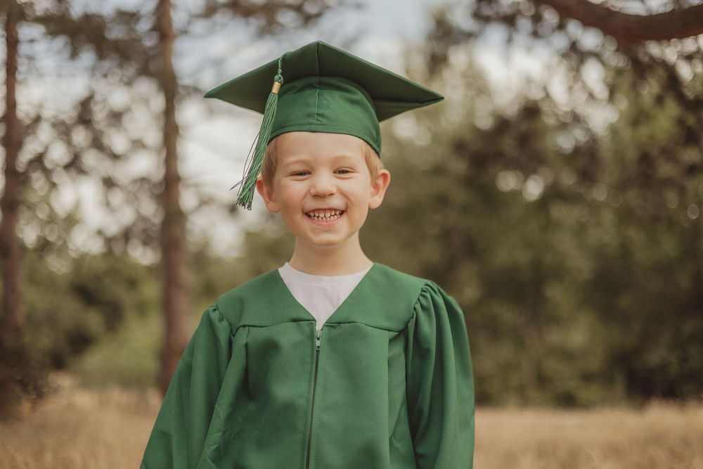 A graduate in green cap and gown smiles brightly during an outdoor photo session in a natural setting.