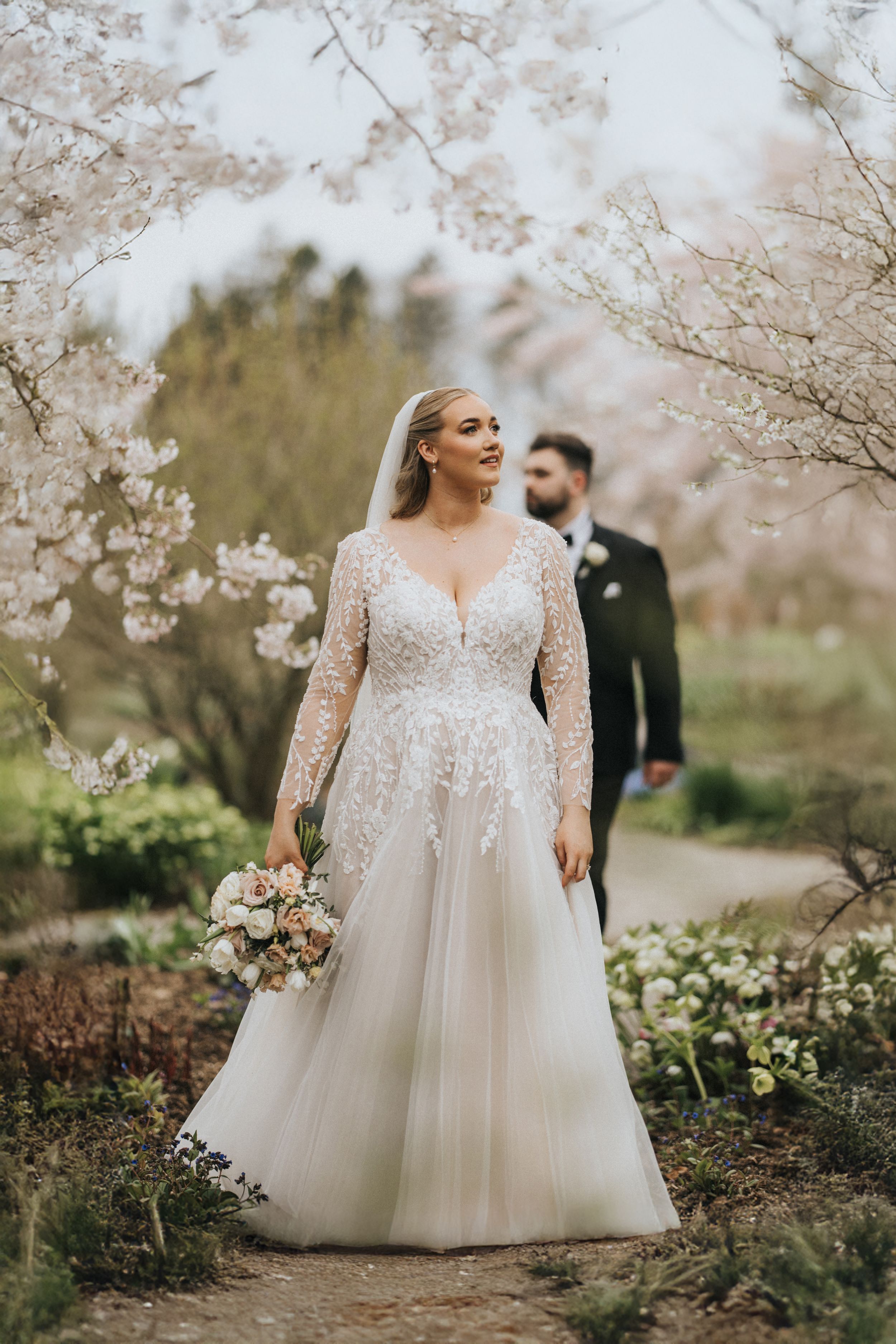 Elegant spring wedding photo with cherry blossoms framing a bride in a lace long-sleeved gown holding a bouquet.