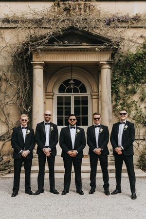 Five groomsmen in black tuxedos and sunglasses pose in front of an elegant doorway with climbing vines.