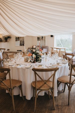 Elegant wedding reception table with white linens, rustic wooden cross-back chairs and draped fabric ceiling in a tent.