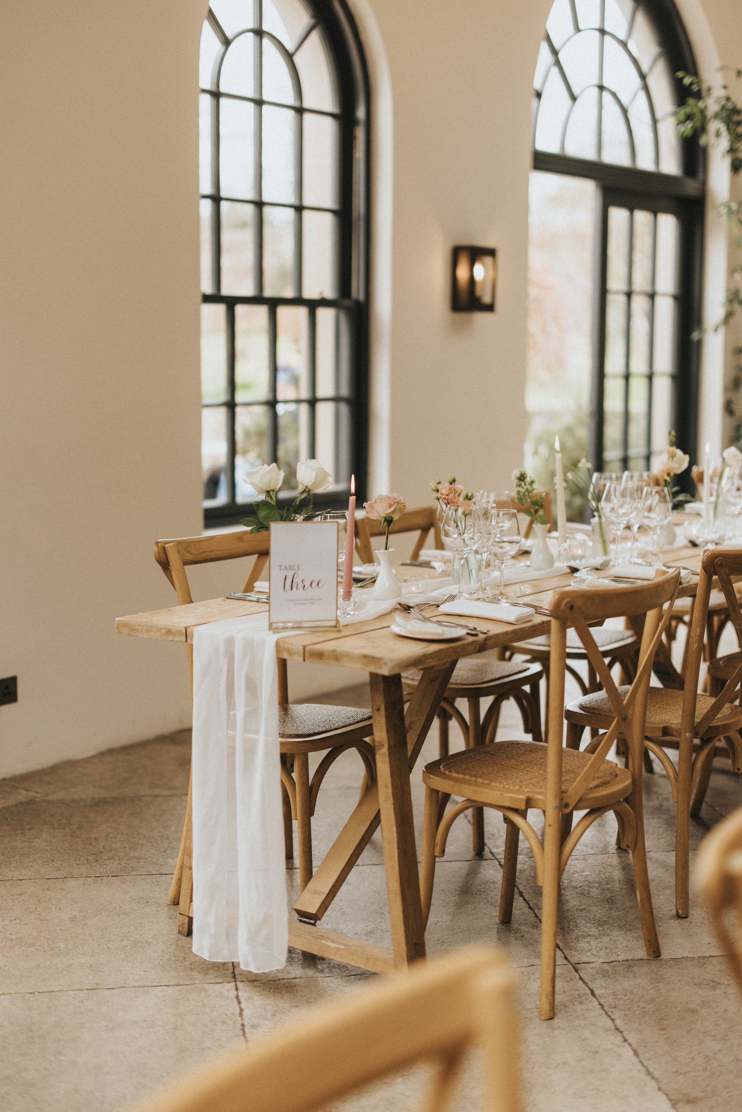 Elegant wedding reception table setup with wooden crossback chairs and tall arched windows in rustic venue setting.