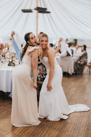 Two women in light-colored formal dresses dancing and celebrating at a wedding reception under white tent decor.