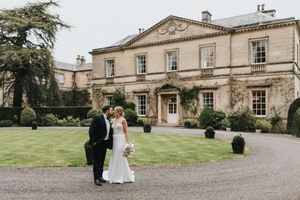A couple on their wedding day stands on the driveway in front of a grand English manor house with manicured gardens.