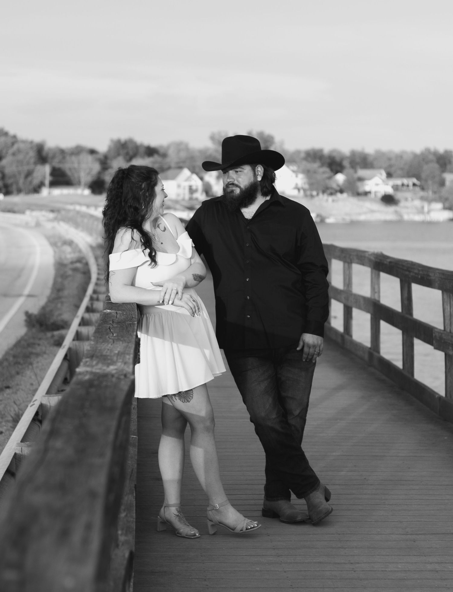A couple in black and white stand together on a wooden bridge walkway with a scenic waterfront view in the background.