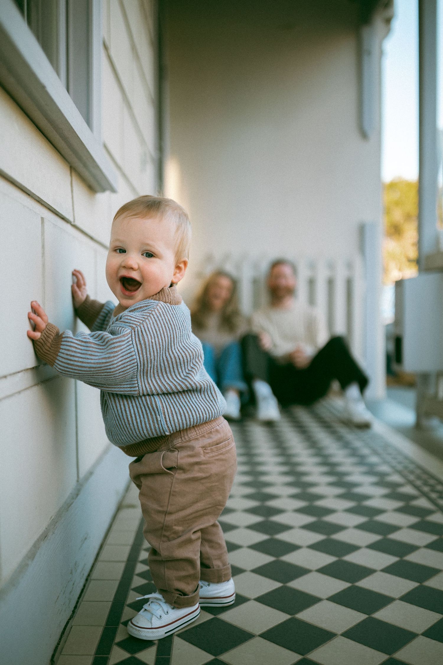 A small child stands smiling on a checkered floor porch in Richmond with his parents in the background.