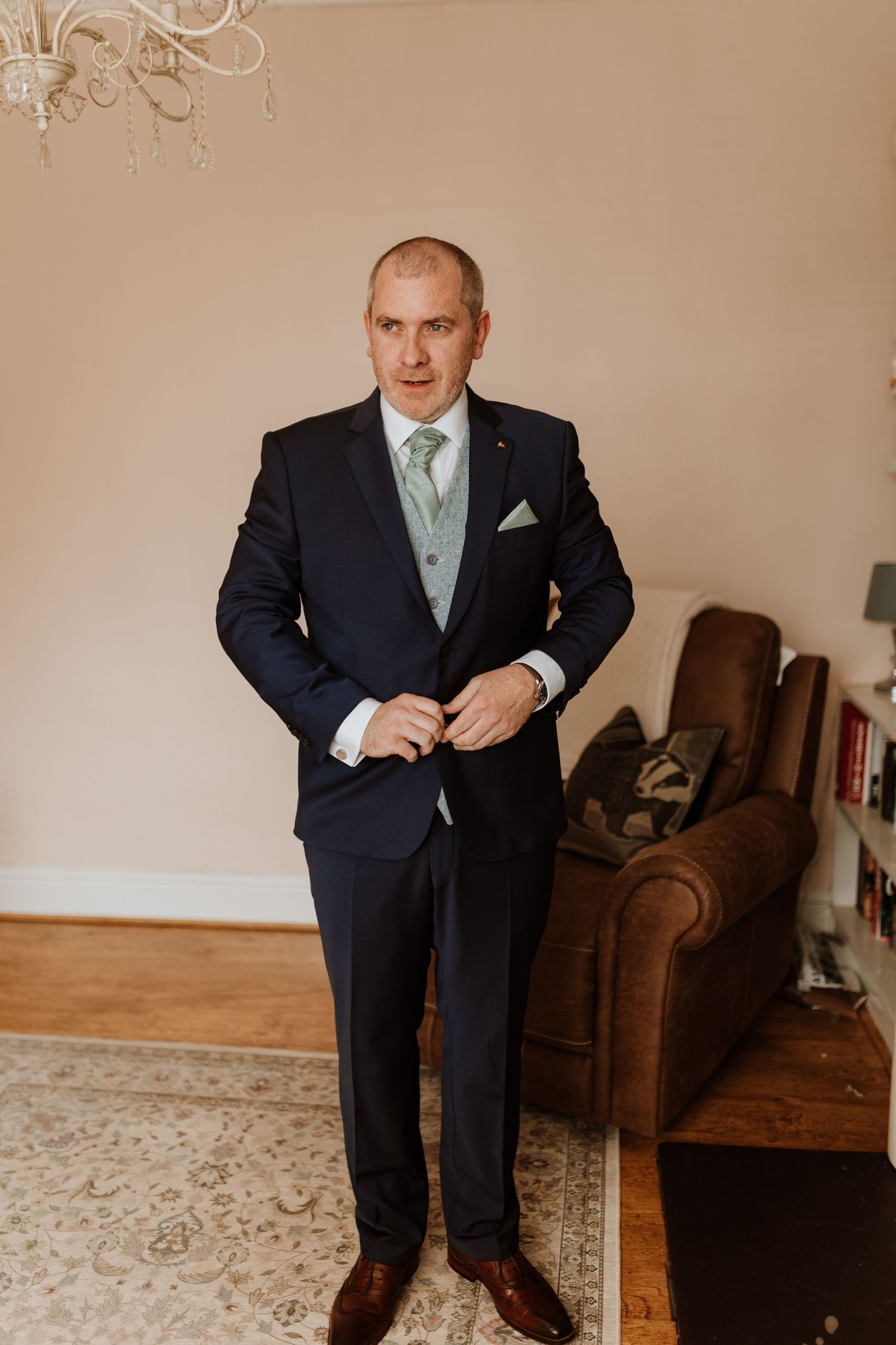 Groom in a suit with a mint green tie stands formally in a room with a leather armchair and hardwood floor.