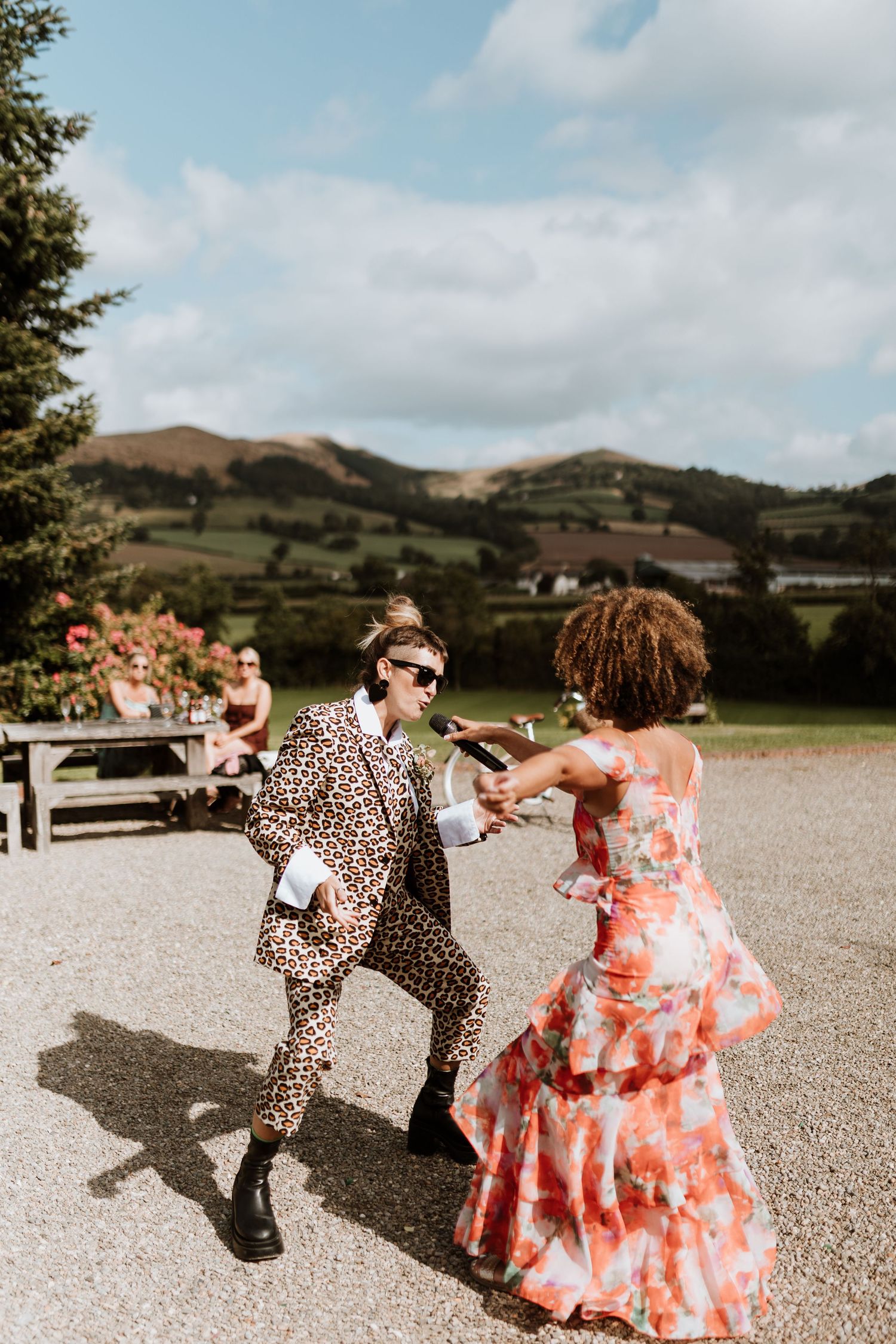 People dancing joyfully on a gravel surface with scenic rolling hills visible in the background.