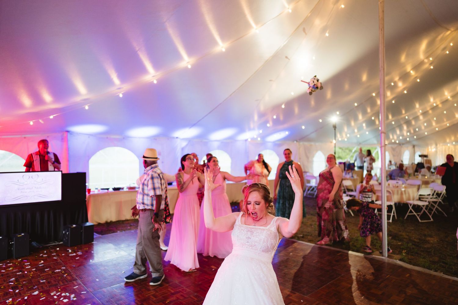 Wedding reception party inside a white tent with dance floor and colorful lighting showing guests celebrating.