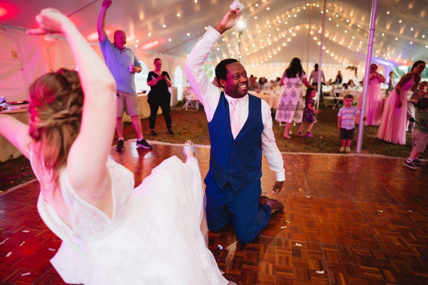 Wedding reception dance party in a tent with twinkling lights and guests dancing energetically on the wooden dance floor.