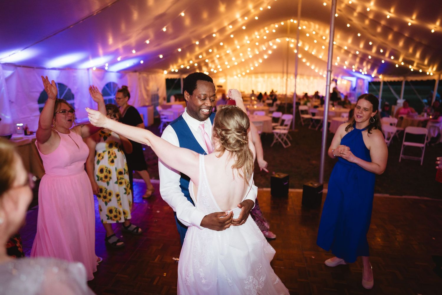Newlyweds dance under twinkling lights at an evening wedding reception with guests in pink and blue dresses watching.
