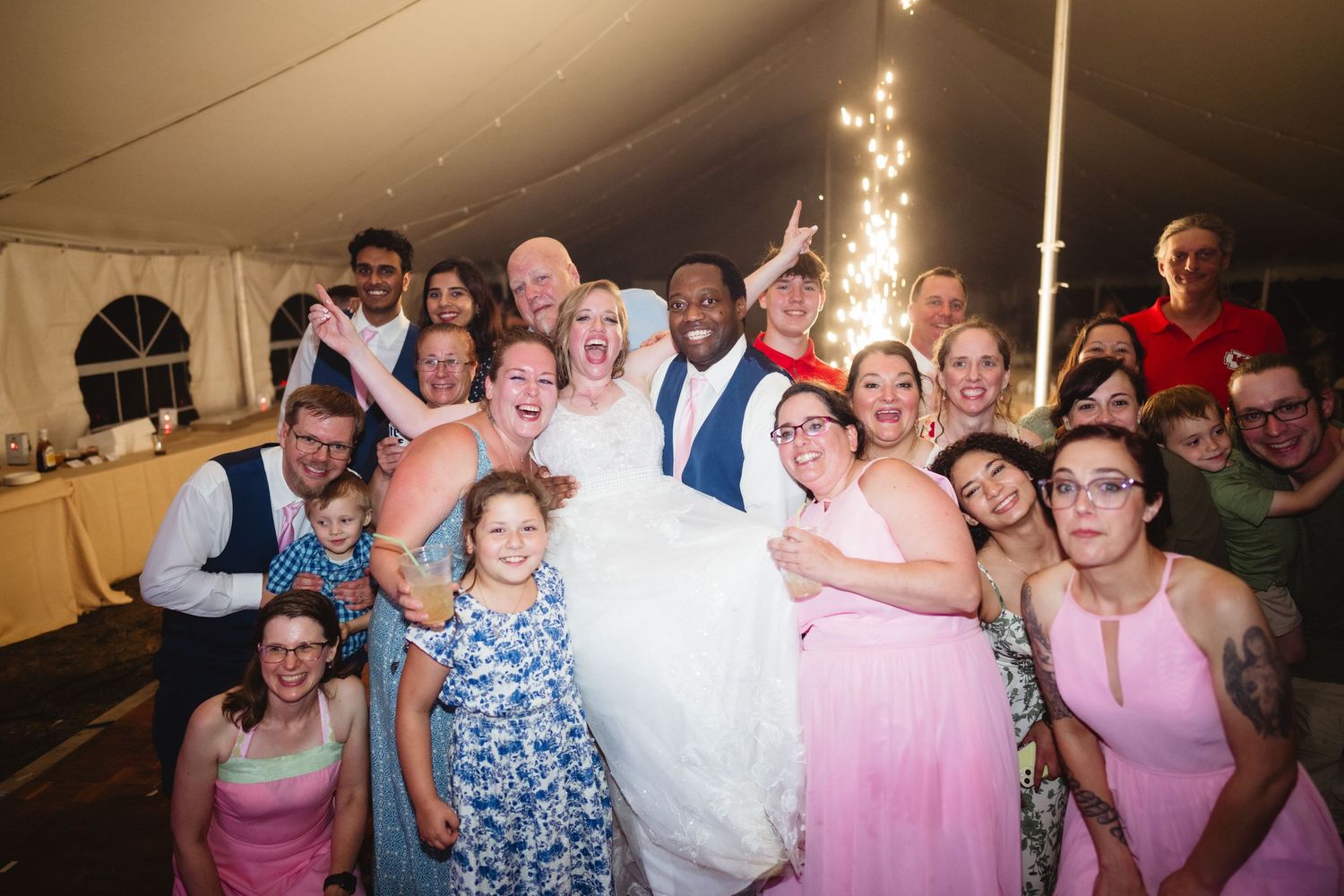 Wedding guests dance and laugh together during a reception under a white tent with glowing lights.