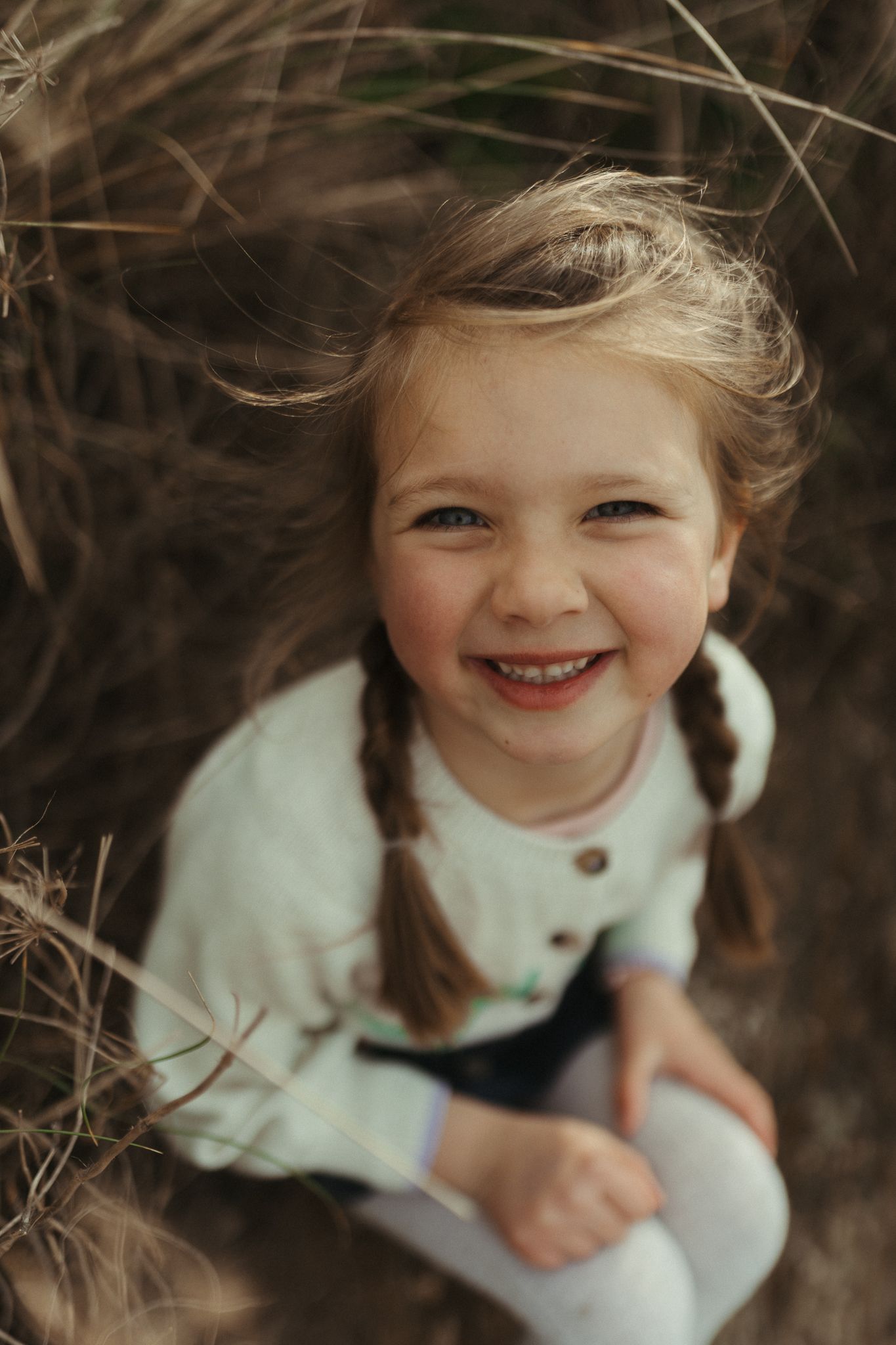 A child in a white cardigan sits surrounded by tall grass while giving a bright smile.