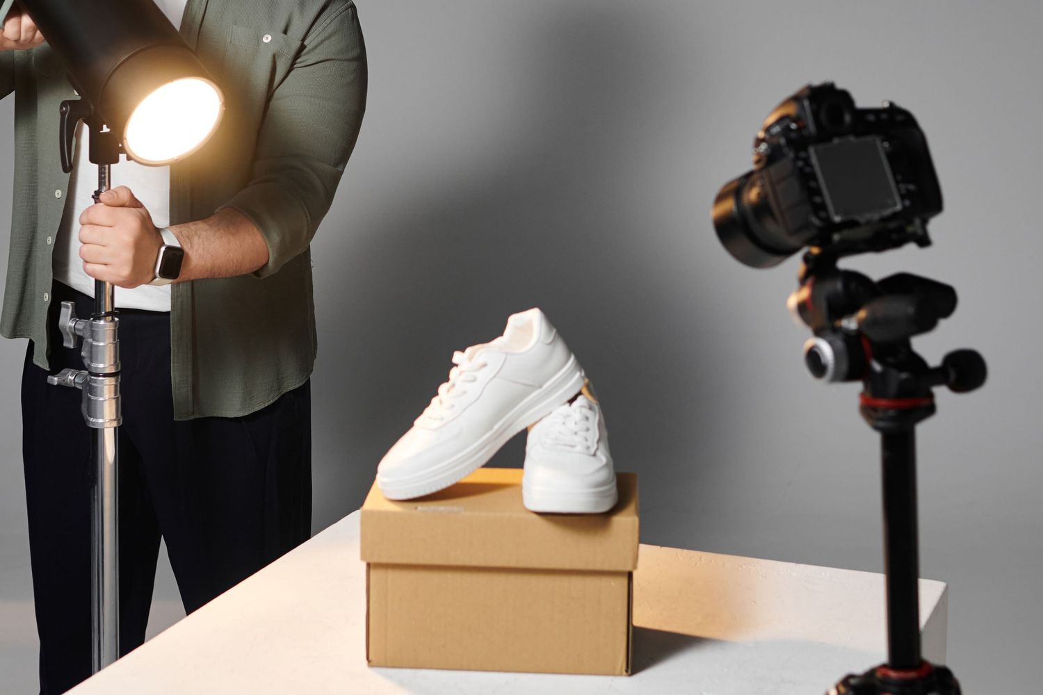 Product photography setup with white sneakers on brown box, camera on tripod, and lighting equipment in studio setting.