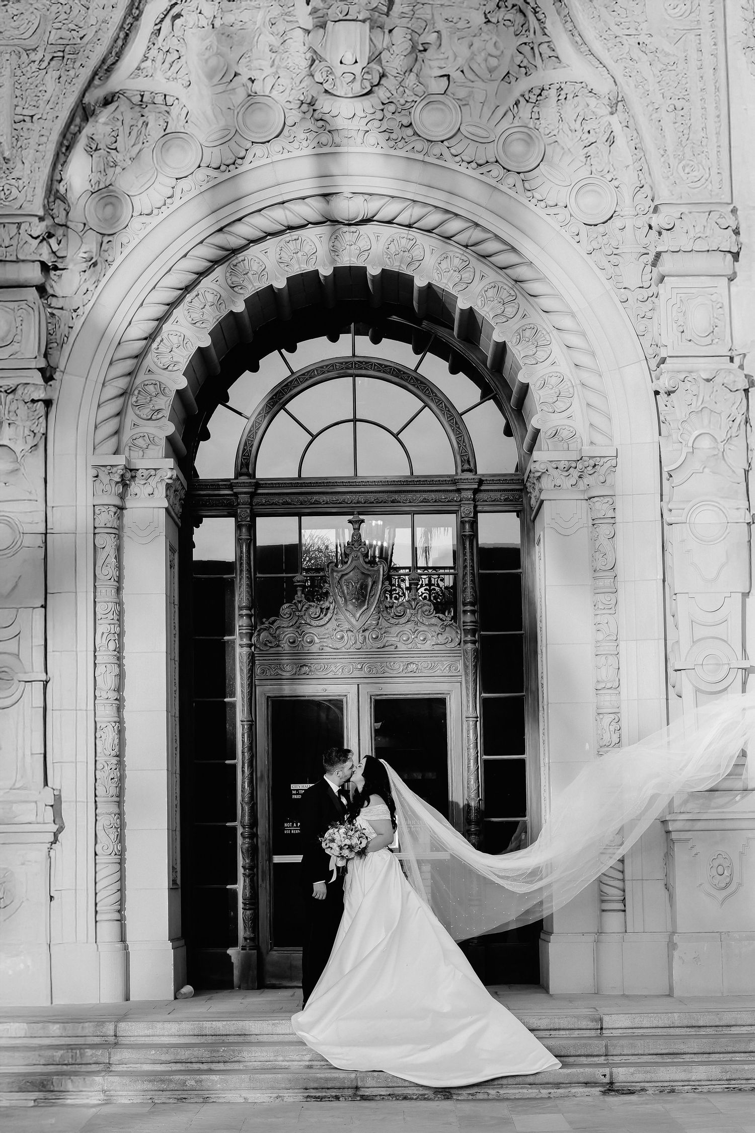 A bridal wedding photograph captured in black and white under an ornate arched doorway with flowing veil.