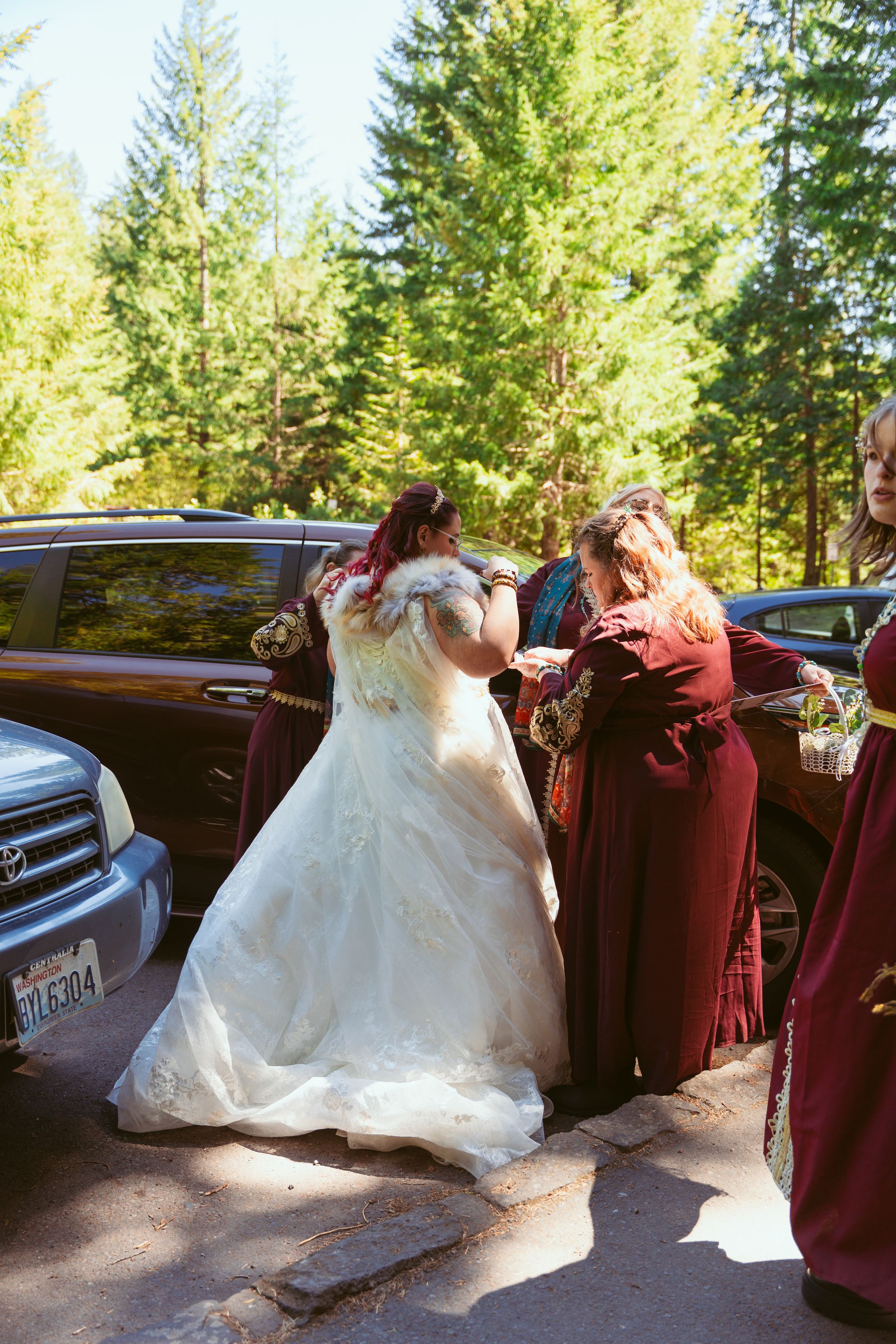 Wedding party members in burgundy dresses helping with a white wedding gown outdoors near parked cars.