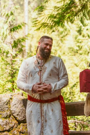 A person in traditional white embroidered clothing stands in a sunlit forest setting with decorative red accents.