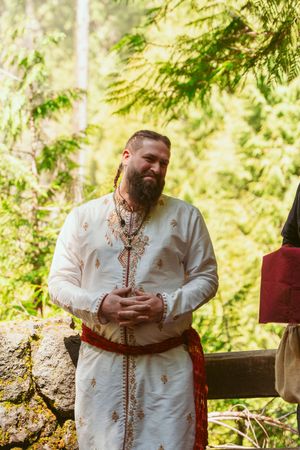 Person in traditional white wedding attire with red sash standing in a forest clearing.