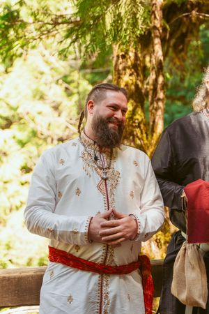 Man in ornate white medieval garments with red sash smiles during outdoor ceremony.