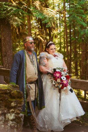 A wedding couple poses in a forest setting with medieval-inspired attire and a vibrant red bouquet.