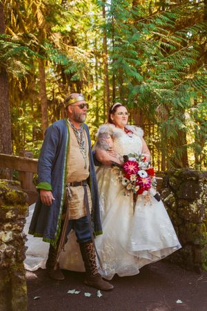 A couple in medieval-inspired wedding attire poses on a forest path holding a vibrant bouquet.