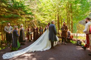 A dreamy forest wedding ceremony takes place on a path surrounded by tall evergreen trees and sunlit foliage.