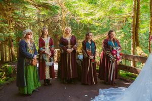 Wedding attendants in medieval-style dresses stand in formation during an outdoor forest ceremony.