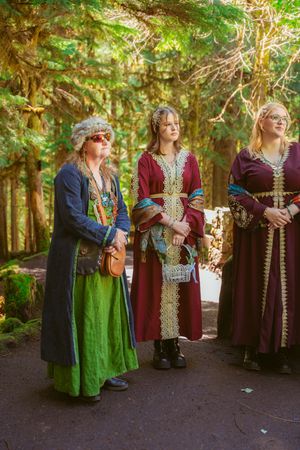 A group in medieval-style costumes with green, burgundy and ornate gold trim stands together in a sunlit forest setting.